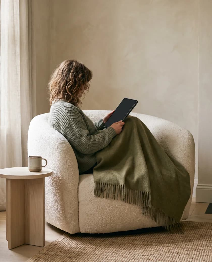 Curled into an armchair under a wool throw, reading from a tablet in late morning light